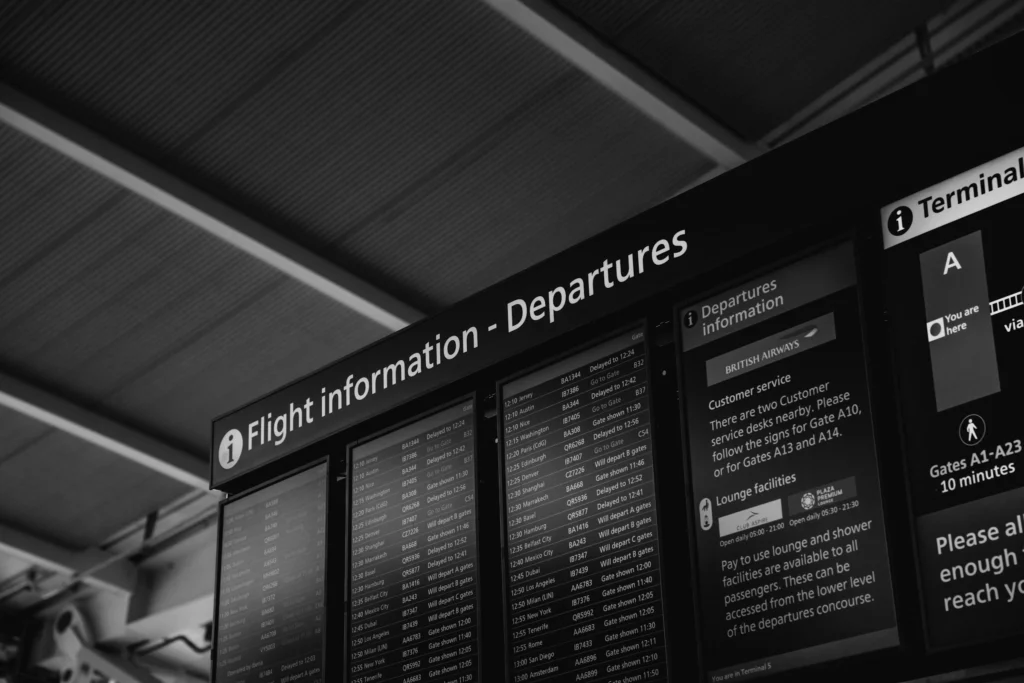 Painel eletrônico exibindo horários de partidas de voos no saguão do aeroporto com passageiros ao fundo.